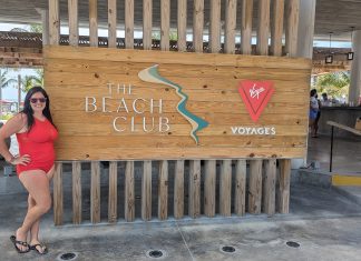 woman standing next to a sign that says "the beach club". She smiling enjoying her adult-only cruise