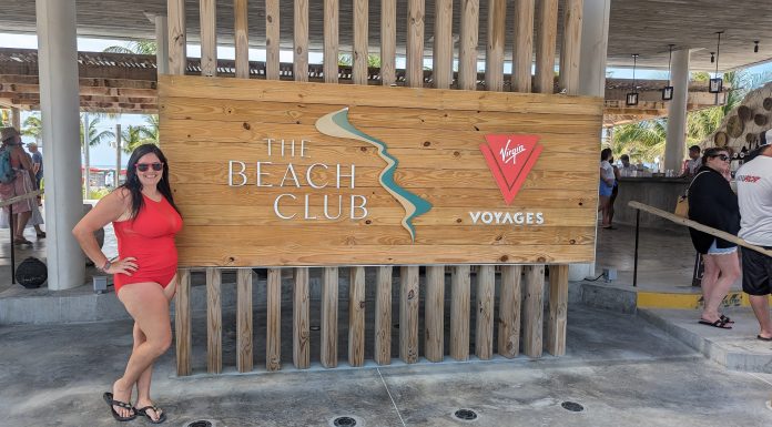 woman standing next to a sign that says "the beach club". She smiling enjoying her adult-only cruise