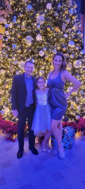 a family (dad, young girl, mom) smiling at the camera in front of a large tree on a cruise; cruising over the holidays