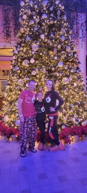 a family (dad, young girl, and mom) smiling at the camera in front of a large tree on a cruise; cruising over the holidays