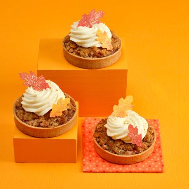 three apple tarts with frosting and leaves on top all set on orange risers with orange background