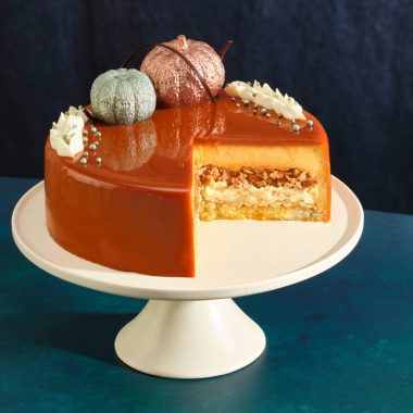 orange cake with big slice missing and pumpkins on top on a white cake stand on a blue and teal background