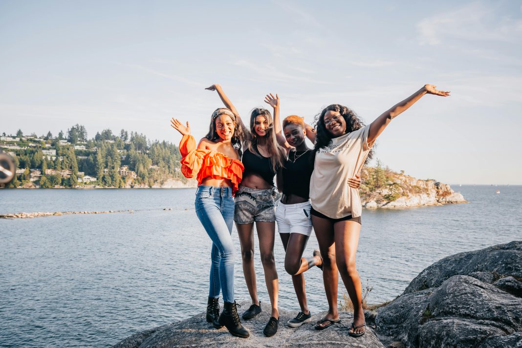 four women laughing and smiling at the camera on a rock in front of water.