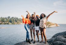 four women laughing and smiling at the camera on a rock in front of water.