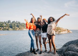 four women laughing and smiling at the camera on a rock in front of water.