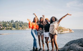 four women laughing and smiling at the camera on a rock in front of water.