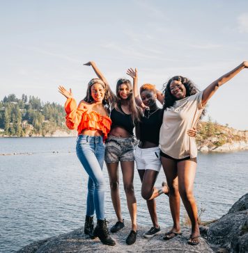 four women laughing and smiling at the camera on a rock in front of water.