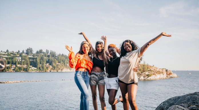 four women laughing and smiling at the camera on a rock in front of water.