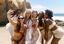 six women smiling together taking a selfie on the beach for their self-care getaway