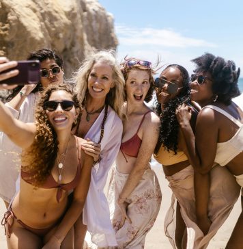 six women smiling together taking a selfie on the beach for their self-care getaway