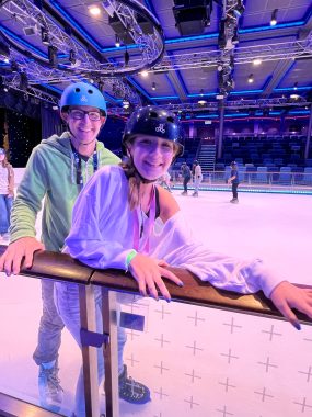 father and daughter with helmets on, smiling at camera, holding onto a rail because they're ice skating; cruising with teens