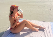 woman dribking coffee and eating a bagel on the beach in her swimwear worried about unexplained weight gain sporting a snackbitches hat