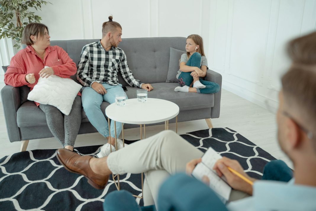 a therapist off to the side looking at a family, mom, dad, and young girl. body language suggests mild tension for family therapy