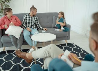 a therapist off to the side looking at a family, mom, dad, and young girl. body language suggests mild tension for family therapy