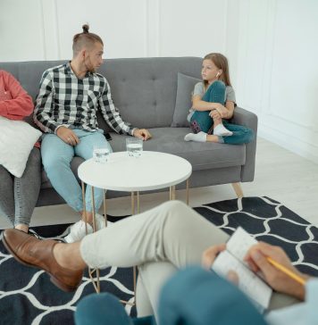 a therapist off to the side looking at a family, mom, dad, and young girl. body language suggests mild tension for family therapy