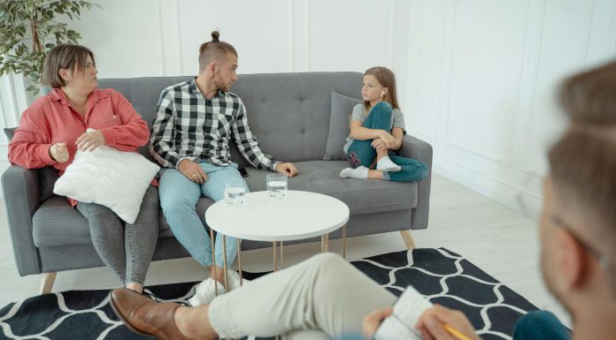 a therapist off to the side looking at a family, mom, dad, and young girl. body language suggests mild tension for family therapy