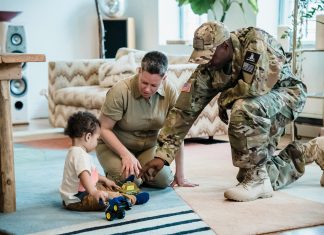 A military mom and dad, both in uniform, help their young child play with toy trucks on a rug in their new living room—capturing a tender moment of family connection during a military moms PCS transition.