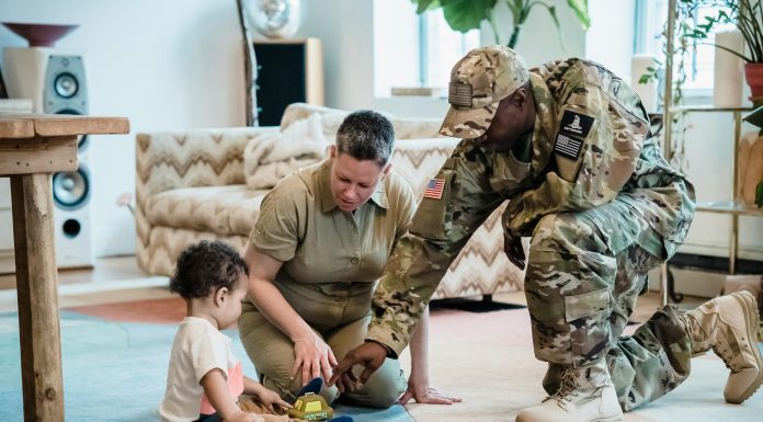A military mom and dad, both in uniform, help their young child play with toy trucks on a rug in their new living room—capturing a tender moment of family connection during a military moms PCS transition.