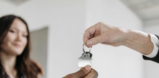 one hand giving another hand a set of keys with a woman smiling in the background, summer real estate tips