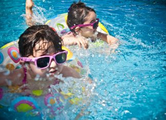 Surviving Summer in Orlando: A Local Mom’s Guide to Beat the Orlando Summer Heat With Kids Two young children wearing sunglasses and colorful swimsuits play in a pool, a fun way of surviving summer in Orlando and staying cool during the heat.