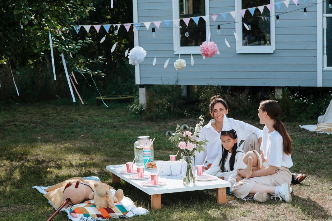pexels-ron-lach-10032942 A joyful outdoor tea party captures a scene of happy childhood, with two women and a young girl smiling together at a low picnic table decorated with pink cups, flowers, and festive bunting.