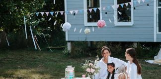 8 Ways to Ensure Your Kids Have a Happy Childhood A joyful outdoor tea party captures a scene of happy childhood, with two women and a young girl smiling together at a low picnic table decorated with pink cups, flowers, and festive bunting.