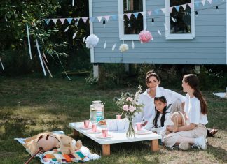 A joyful outdoor tea party captures a scene of happy childhood, with two women and a young girl smiling together at a low picnic table decorated with pink cups, flowers, and festive bunting.