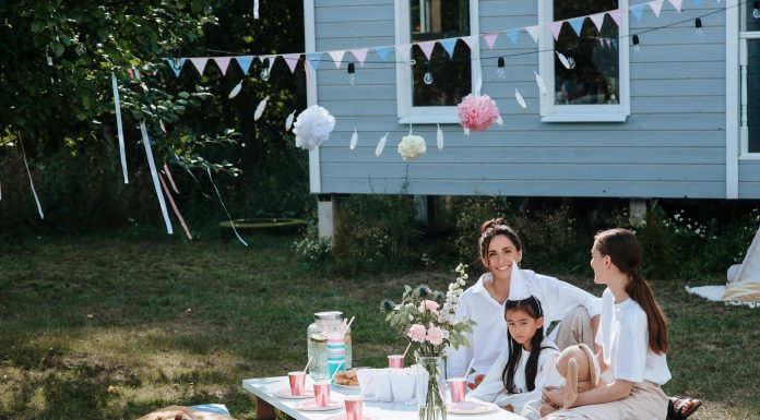 8 Ways to Ensure Your Kids Have a Happy Childhood A joyful outdoor tea party captures a scene of happy childhood, with two women and a young girl smiling together at a low picnic table decorated with pink cups, flowers, and festive bunting.