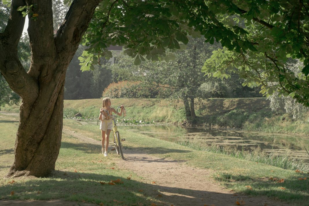 pexels-ron-lach-10601059 Young girl walking her yellow bike along a leafy pond path on a sunny day, illustrating screen-free summer activities for kids.
