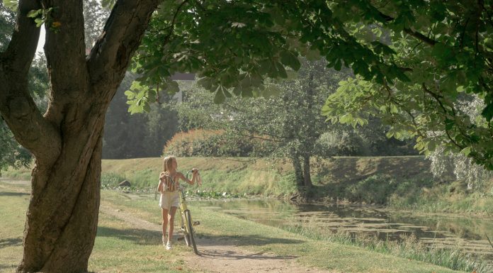 17 Screen-Free Summer Activities for Kids Young girl walking her yellow bike along a leafy pond path on a sunny day, illustrating screen-free summer activities for kids.