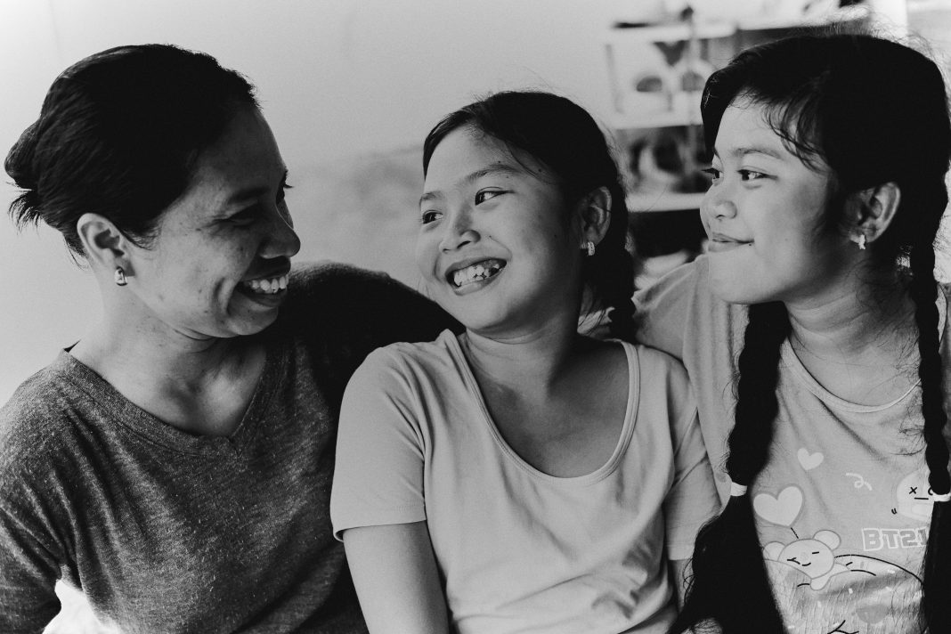 pexels-vikkirillova-33194119 Black and white image of a smiling mom sitting on the floor with her children in a cozy living room, showing a moment of connection and balance, reflecting Mom Back to School health.